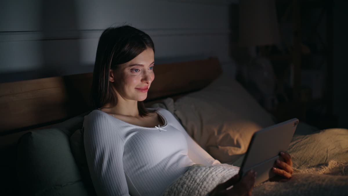 Woman relaxing in bed at night, browsing a website on her tablet with her face lit by the screen